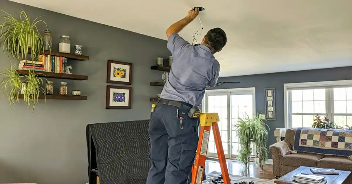 Man on a ladder installing or repairing a ceiling light fixture in a living room with dark gray walls and wooden floating shelves.