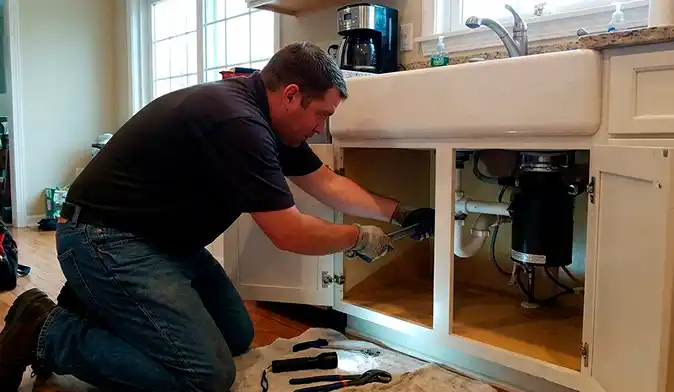 A plumber kneeling on the floor, using a wrench to repair the white PVC drain pipes and garbage disposal unit under a kitchen farmhouse sink.